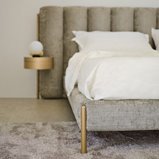 Luxury Modern Bed With Wide Headboard A stylish bedroom showcases the Luxury Modern Bed With Wide Headboard, adorned in white bedding. Adjacent to it is a round, golden nightstand supporting a spherical white lamp. Beneath the bed lies a soft gray rug, enhancing the room's modern elegance.