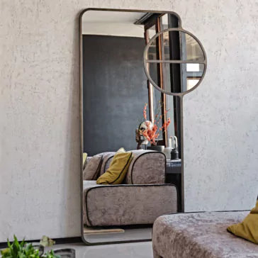 A modern living room with a Luxury Modern Floor Mirror leaning against a white textured wall. The mirror reflects a gray couch adorned with yellow pillows and a vase of orange flowers on a side table. Above the couch is a round window, and in the foreground, there's a potted plant.