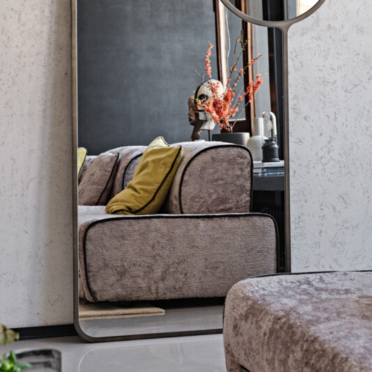 A cozy living room scene is beautifully captured in the reflection of the Luxury Modern Floor Mirror. The mirror reveals a plush taupe sofa adorned with a vibrant yellow pillow, and behind the sofa, a window frames a vase of red dried flowers, enhancing the room's modern, neutral aesthetic.