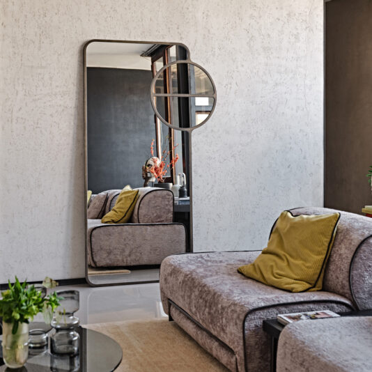 A modern living room with the Luxury Modern Floor Mirror propped against a textured white wall. A light gray sofa with yellow cushions is in the foreground. A small round mirror is hanging above the tall, modern mirror. A coffee table with plants and decor is nearby.