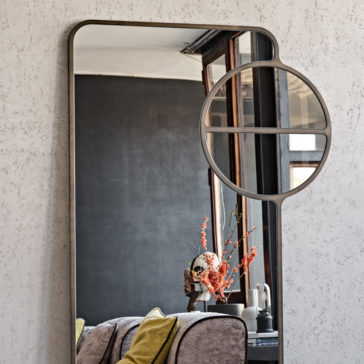 The Luxury Modern Floor Mirror, featuring a circular accent overlay, is mounted on a textured wall. It reflects a cozy living space that includes a sofa adorned with yellow cushions, a table displaying a decorative vase of orange flowers, and a large window letting in natural light.