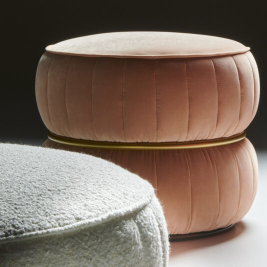 A close-up shot of two Luxury Modern Round Upholstered Pouffes. The one in the background is upholstered in a smooth, dusky pink fabric with pleats and a gold accent, while the one in the foreground is covered in textured, light grey fabric. Both pouffes have a soft, cushioned appearance perfect for any modern decor.