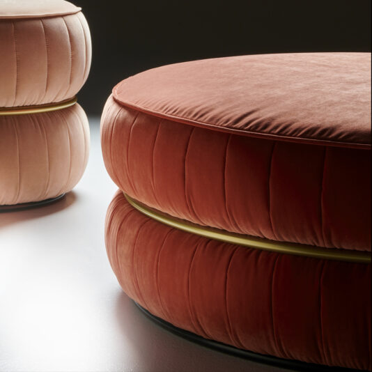 A close-up view of two Luxury Modern Round Upholstered Pouffes, one in a soft peach shade and the other in a rich reddish-orange tone. Both elegant pouffes feature a pleated, cushioned design and are set against a dark background.