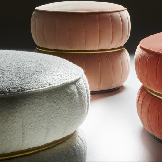 Close-up of stylish, Luxury Modern Round Upholstered Pouffes grouped together. The pouffe in the foreground has a white textured fabric, while the others in the background are upholstered in a soft, pleated peach fabric. Each pouffe features a gold-colored accent ring at the base.