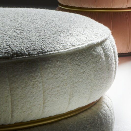 A close-up of two Luxury Modern Round Upholstered Pouffes with metallic bases. The foreground pouffe features a white textured fabric, while the one in the background showcases a smooth light peach finish. Both are set on a reflective surface, accentuating their elegant design details.