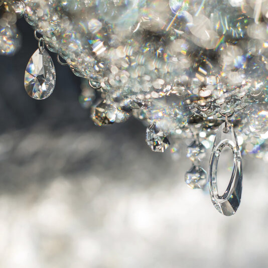 A close-up shot of the Luxury Oval Crystal Chandelier, showcasing sparkling, crystal-clear hanging ornaments that capture light and create a prism effect. Vibrant reflections and bokeh fill the background, highlighting an array of teardrop and oval-shaped crystals.