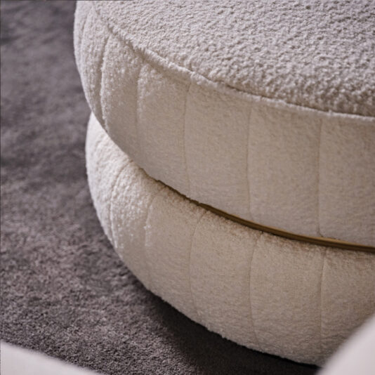 Luxury Round Bouclé Pouffe A close-up of the Luxury Round Bouclé Pouffe, showcasing its textured, cream-colored bouclé fabric and soft, plush surface. The circular ottoman features a distinctive layered, stacked design and is placed on a grey carpeted floor.
