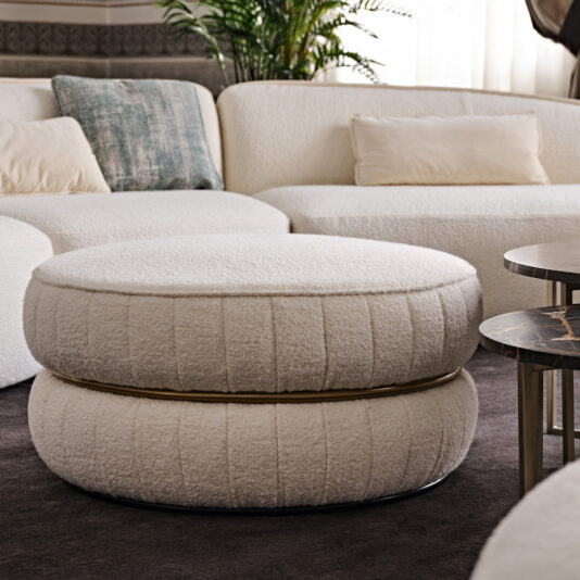 A contemporary living room showcases white, rounded cushioned seats along with a large, plush, circular ottoman featuring a textured surface. Positioned behind the Luxury Round Bouclé Pouffe are matching curved white couches embellished with light-colored pillows. A potted plant in the background provides a natural touch.