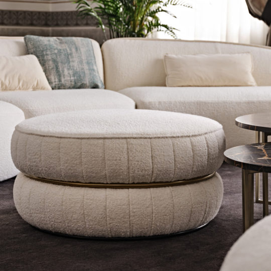 A contemporary living room showcases white, rounded cushioned seats along with a large, plush, circular ottoman featuring a textured surface. Positioned behind the Luxury Round Bouclé Pouffe are matching curved white couches embellished with light-colored pillows. A potted plant in the background provides a natural touch.