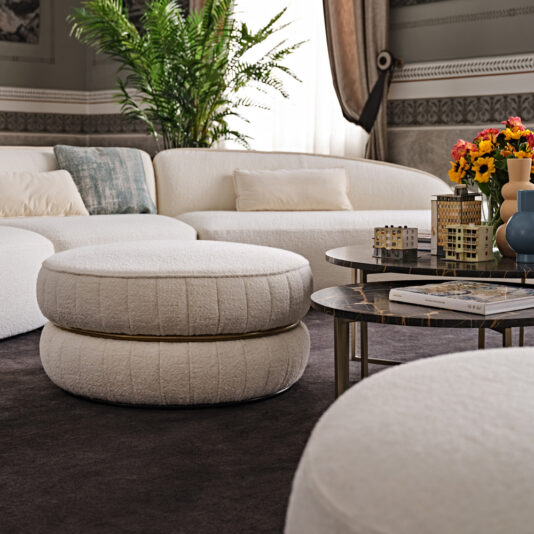 Luxury Round Bouclé Pouffe A modern living room showcases a white, curved sofa paired with a Luxury Round Bouclé Pouffe. A dark round coffee table adorned with decorative items is centered on a dark carpet. A potted green plant and a bouquet of flowers in a blue vase infuse color into the neutral, luxury decor.