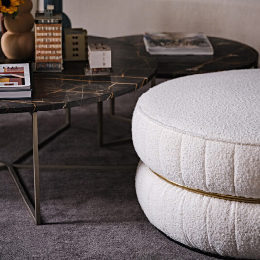 Luxury Round Bouclé Pouffe A cozy living room setup featuring a modern coffee table with a dark marble top, showcasing miniature building models and a stack of books. Adjacent to it are two Luxury Round Bouclé Pouffes, stacked on top of each other, adding texture and elegance to the gray carpet.