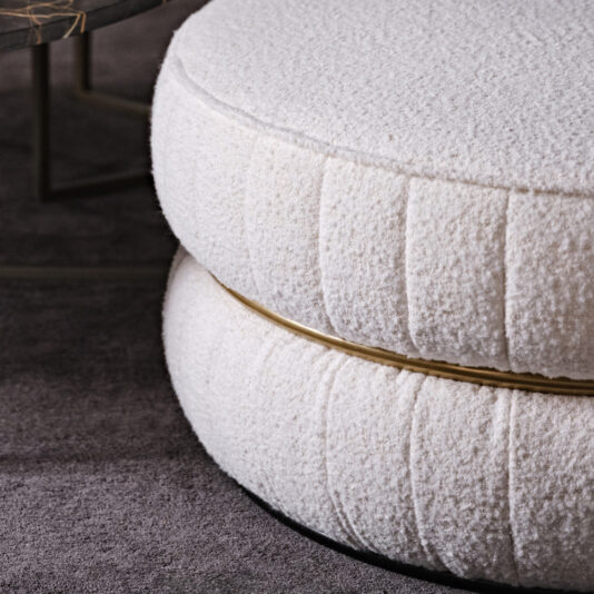 Luxury Round Bouclé Pouffe Close-up of two Luxury Round Bouclé Pouffes with white tufted upholstery, stacked on top of each other and separated by a golden band. The textured and fuzzy material gives these elegant ottomans a refined touch. A portion of a dark modern coffee table can be seen in the background on a dark carpeted floor.