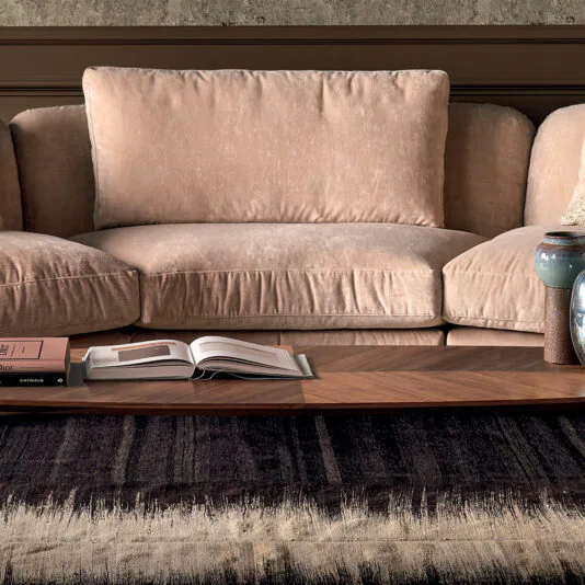 A Luxury Velvet Modular Sofa with plush cushions is positioned against a wall. In front of the sofa, a wooden coffee table holds an open book, a few closed books, and a ceramic mug. A dark, textured area rug extends from under the coffee table.