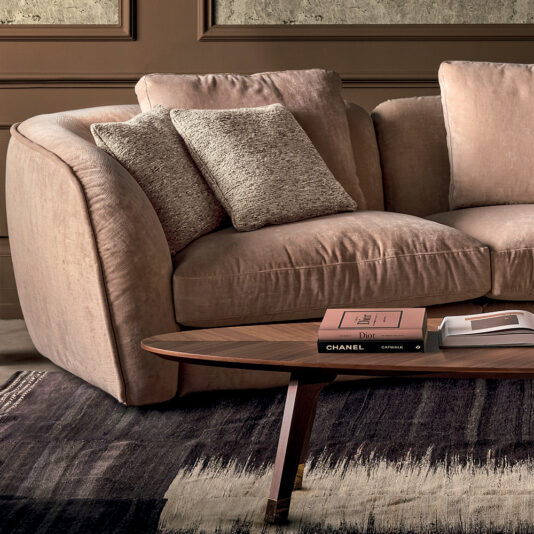 A cozy Luxury Velvet Modular Sofa in beige, adorned with plush cushions, is positioned against an earth-toned wall. In front, a wooden coffee table displays a book titled 