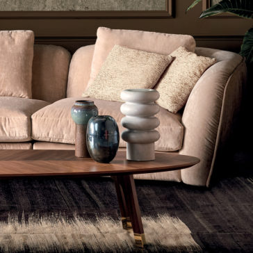 Luxury Velvet Modular Sofa A cozy living room featuring the Luxury Velvet Modular Sofa adorned with two textured pillows. In front of the sofa, there's a wooden coffee table displaying three decorative vases in various shapes and colors. A large leafy plant can be partially seen in the background.