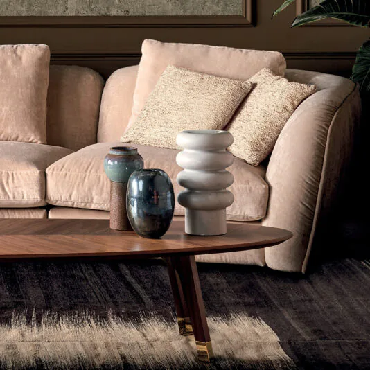 A cozy living room featuring the Luxury Velvet Modular Sofa adorned with two textured pillows. In front of the sofa, there's a wooden coffee table displaying three decorative vases in various shapes and colors. A large leafy plant can be partially seen in the background.