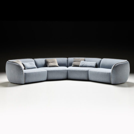 The Modern Bouclè Modular Corner Sofa, featuring a light blue hue and rounded edges, is elegantly placed against a black backdrop. This L-shaped sectional comes with grey and beige accent pillows, presenting a contemporary and cozy look ideal for a spacious living room.