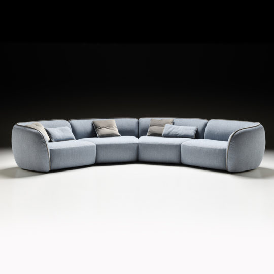 The Modern Bouclè Modular Corner Sofa, featuring a light blue hue and rounded edges, is elegantly placed against a black backdrop. This L-shaped sectional comes with grey and beige accent pillows, presenting a contemporary and cozy look ideal for a spacious living room.