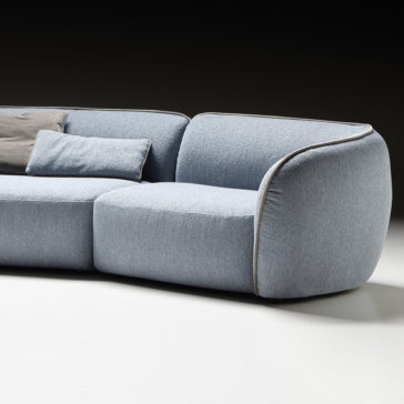Modern Bouclè Modular Corner Sofa The Modern Bouclè Modular Corner Sofa, upholstered in blue and set against a dark background, is featured prominently. The sofa showcases clean lines, plush cushions, and a minimalist design. A couple of gray throw pillows are placed on the seat, providing a touch of contrast to the overall aesthetic.