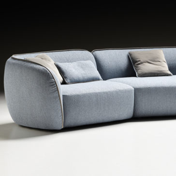 Modern Bouclè Modular Corner Sofa The Modern Bouclè Modular Corner Sofa in light gray, boasting a minimalist design, is positioned against a dark background. This sectional showcases smooth, curved edges and comes with two gray cushions and a smaller rectangular pillow. Its textured upholstery gives it a soft-to-the-touch appearance.