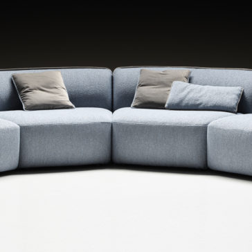 Modern Bouclè Modular Corner Sofa A Modern Bouclè Modular Corner Sofa with light blue upholstery is positioned against a dark background. It features three gray throw pillows neatly arranged on the seats, creating a stylish and comfortable appearance. The flooring is a light neutral color.