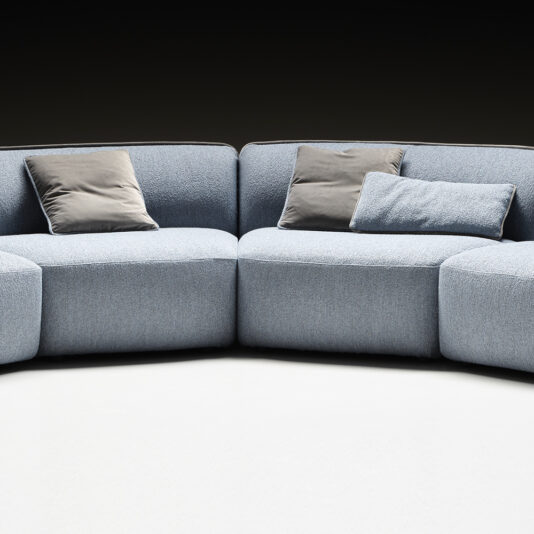 A Modern Bouclè Modular Corner Sofa with light blue upholstery is positioned against a dark background. It features three gray throw pillows neatly arranged on the seats, creating a stylish and comfortable appearance. The flooring is a light neutral color.