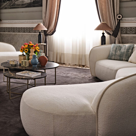 A luxurious living room with a Modern Chaise Style Bouclé Modular Sofa and a dark round coffee table in the center, adorned with vibrant flowers and decor pieces. The space features elegant gray and white patterned wallpaper, a plush dark gray rug, and sheer curtains allowing soft light in.