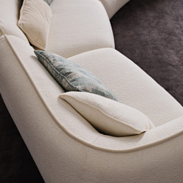 A top view of the Modern Chaise Style Bouclé Modular Sofa, featuring plush cushions and arranged in a curved layout. The sofa boasts bouclé textured upholstery and includes two cushions: one beige and one pale blue with a subtle pattern. The background highlights a dark carpeted floor.