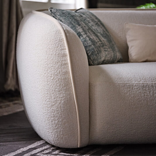 Close-up of the Modern Chaise Style Bouclé Modular Sofa in white with a textured fabric and a rounded armrest. The sofa is decorated with two throw pillows—one featuring a muted blue pattern and the other in solid beige. The setting includes a patterned rug and draped curtains in the background.