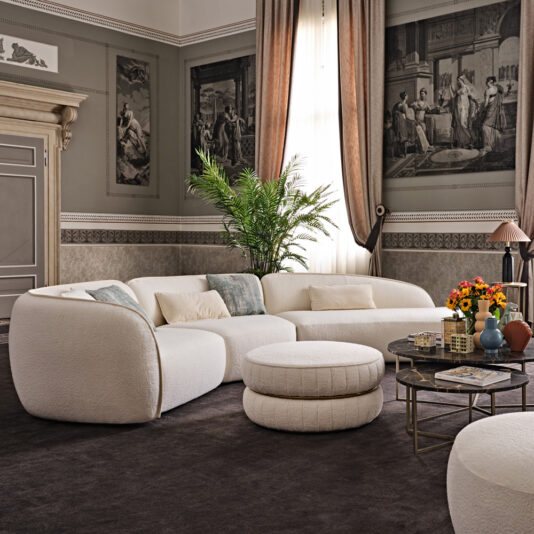 A sophisticated living room boasts a Modern Chaise Style Bouclé Modular Sofa in elegant white, complemented by matching round ottomans. The space is enriched by a dark carpet, classical wall art, tall drapes, a potted plant, and a coffee table adorned with books and a colorful flower arrangement.
