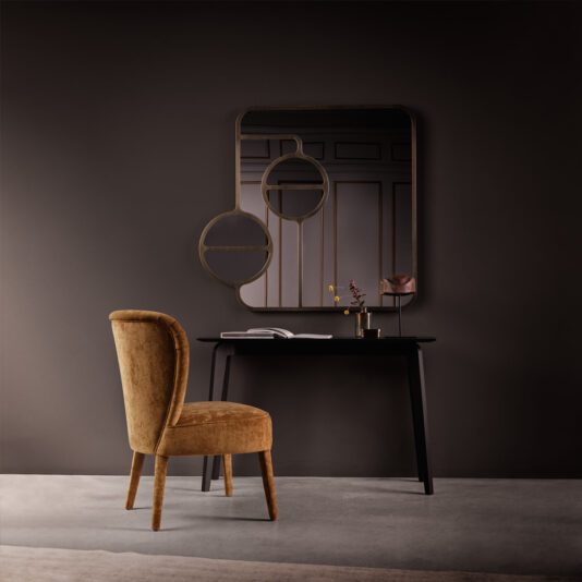 The image showcases a stylish and contemporary interior setting, featuring a mustard-colored upholstered chair positioned in front of a sleek black console table. The table holds an open book, a small vase with pink flowers, and a decorative sculpture. Mounted on the dark, textured wall behind the table is the Modern Designer Curved Bronze Wall Mirror. A light grey rug covers the floor.