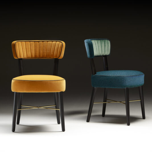Two elegant, contemporary chairs placed side by side. The Modern Designer Dining Chair on the left features a mustard-yellow velvet seat and backrest, while the chair on the right has a teal-blue velvet backrest and a dark blue fabric seat. Both have black legs with gold accents.