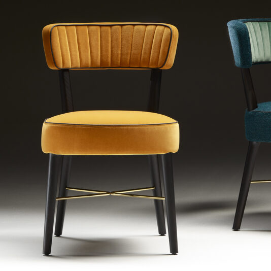 Modern Designer Dining Chair The Modern Designer Dining Chair boasts a mustard yellow, upholstered seat and backrest with sophisticated vertical stitching on the backrest. It features sleek black metal legs connected by an elegant brass crossbar. Additionally, part of another similar chair in a vibrant teal is partially visible on the right side.