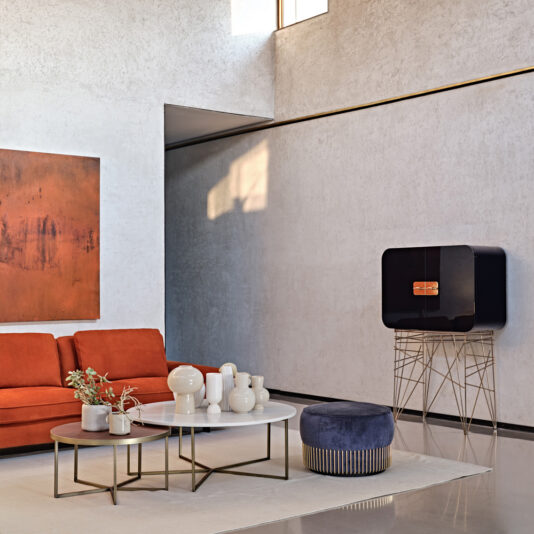 In a modern living room, a burnt orange couch stands out along with a large abstract painting on the wall. A minimalist TV stand holds up a black television, while a Modern Lacquered Cocktail Bar Cabinet adds an elegant touch beside the centerpiece coffee table adorned with white vases of varying shapes. Natural light streams in from a high window.