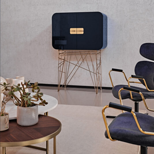 A chic, modern living room featuring the Modern Lacquered Cocktail Bar Cabinet with golden accents, supported by geometric gold legs. Accompanying the cabinet is a white round coffee table with a potted plant, and cozy chairs with gold frames and dark cushions.
