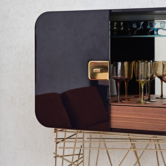 The Modern Lacquered Cocktail Bar Cabinet, featuring a sleek glossy black door that is partially open, reveals an array of vibrant wine glasses inside. The cabinet is elevated on sophisticated gold-toned geometric metal legs, with the reflection of a nearby sofa visible on the door's polished surface.