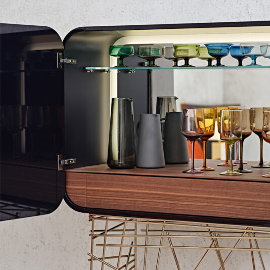 The Modern Lacquered Cocktail Bar Cabinet boasts a stylish, modern design. When you open the door, it reveals a wooden shelf holding colorful wine glasses, a couple of pitchers, and additional glassware. The cabinet features a reflective interior and is softly illuminated for an elegant touch.