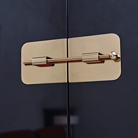 The Modern Lacquered Cocktail Bar Cabinet features a sleek, polished brass door latch that securely fastens its dark, smooth cabinet doors. Its minimalist design with rounded rectangular plates and a cylindrical bar creates a sophisticated and stylish look against the high-gloss black surface.