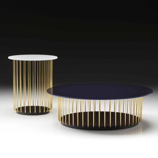 Contemporary Round Glass Coffee Table A pair of modern tables feature vertical gold rods forming the bases—one taller with a small, round white top and the other shorter and wider with a large, round black top. The aesthetic is reminiscent of the Contemporary Round Glass Coffee Table style. Both tables boast minimalist designs and are set against a dark backdrop.