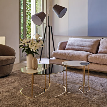 A modern living room showcases a beige sofa and a gray upholstered chair. The Modern Nest Of Coffee Tables, adorned with a vase of white lilies, sits atop a plush beige rug. A chic floor lamp with double black lampshades stands in the corner, near the large windows.