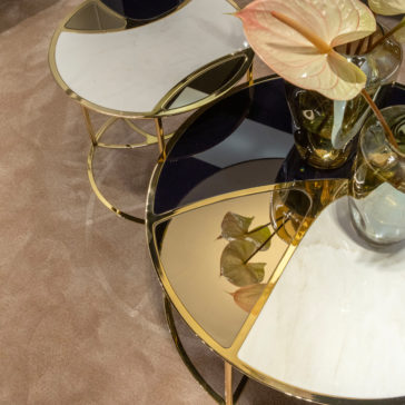 A close-up view of the Modern Nest Of Coffee Tables showcases their elegant marble tops and gold metallic frames. The tables are artistically arranged in a semi-overlapping manner, reflecting a mirrored surface. On one table, a vase containing light pink anthurium flowers is placed.