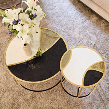 On a plush carpet, the Modern Nest Of Coffee Tables showcases two modern tables. The larger table features a round design with segmented white, beige, and black surfaces, adorned with a vase of white lilies. The smaller table mirrors this minimalist elegance with matching white and black segments. This modern ensemble adds chic sophistication to any space.