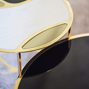 A close-up shot of the Modern Nest Of Coffee Tables shows a sleek design with rounded edges. One table has a white top adorned with gold geometric accents and is supported by gold legs, while its companion features a black top paired with matching gold legs. The white-topped table is slightly elevated and overlaps the black-topped one, creating an elegant and modern aesthetic.