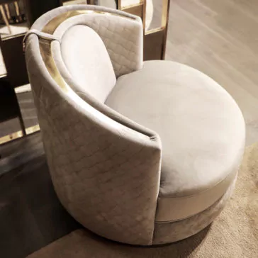 A luxurious, circular Modern Quilted Velvet Swivel Tub Chair features light gray fabric and a plush cushion. Its curved backrest creates an enclosed, cozy seating area, accentuated by a metallic detail running along the top of the backrest. The chair is placed on a soft beige rug that rests on a wooden floor.