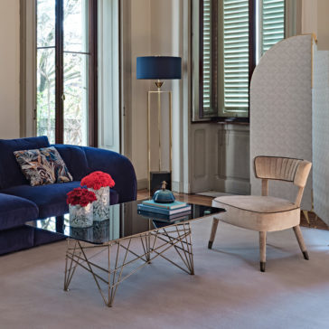 A modern living room features a blue velvet sofa with patterned cushions, a beige armchair, and a blue-shaded floor lamp. The Modern Rectangular Glass Coffee Table with a geometric gold frame holds books and red flowers. A window with green shutters and a decorative screen adorn the background.