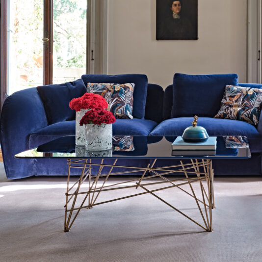 A modern living room featuring a deep blue, plush sofa adorned with patterned cushions. In front of it stands the Modern Rectangular Glass Coffee Table, featuring a geometric gold metal base and holding a vase of red flowers, a blue decorative item, and two books. A painting hangs above the sofa.