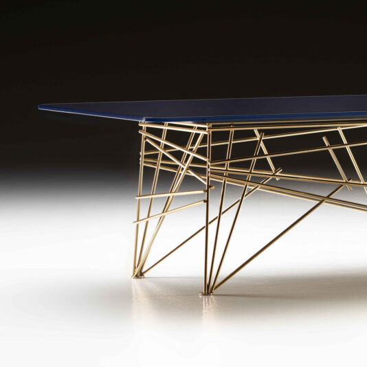 The Modern Rectangular Glass Coffee Table boasts a sleek, dark blue glass top complemented by a distinctive gold metal base, intricately designed with intersecting rods in a geometric pattern. This stylish piece is showcased against a gradient black-and-white background.