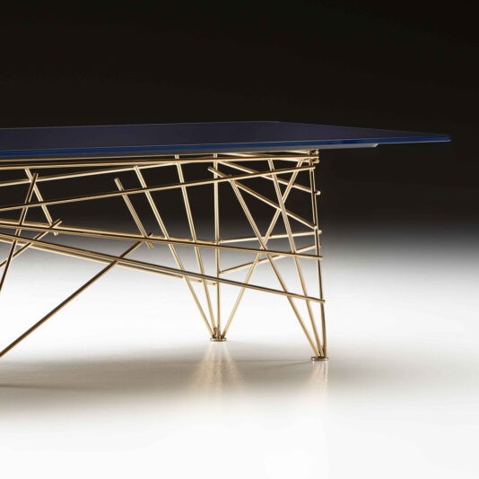 The Modern Rectangular Glass Coffee Table features a blue rectangular top and an intricate geometric gold metal base. Set against a dark background, the illuminated table stands out with its contemporary and artistic design.