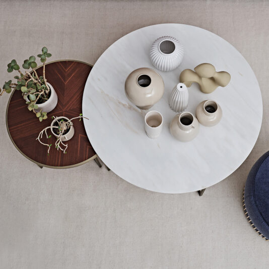 The Modern Set Of Round Coffee Tables, featuring one larger table with a white surface showcasing several neutral-toned ceramic vases and a smaller table with a dark wooden top supporting a couple of potted plants, are elegantly placed on a beige carpet. A blue cushion or stool can be seen partially nearby.