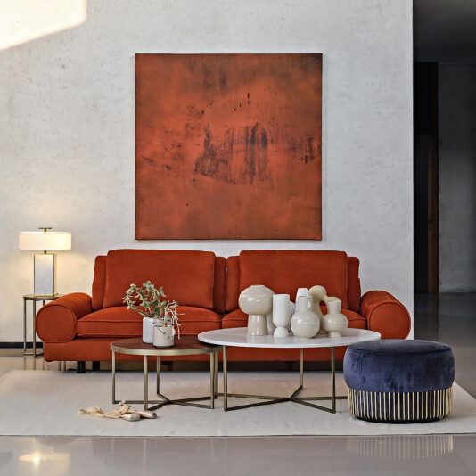 A modern living room showcasing an orange sofa accompanied by a stylish Modern Set Of Round Coffee Tables in front. The tables are adorned with various ceramic vases and objects. Above the sofa hangs a large abstract painting with red hues. A blue ottoman and a lamp on a side table add to the decor.